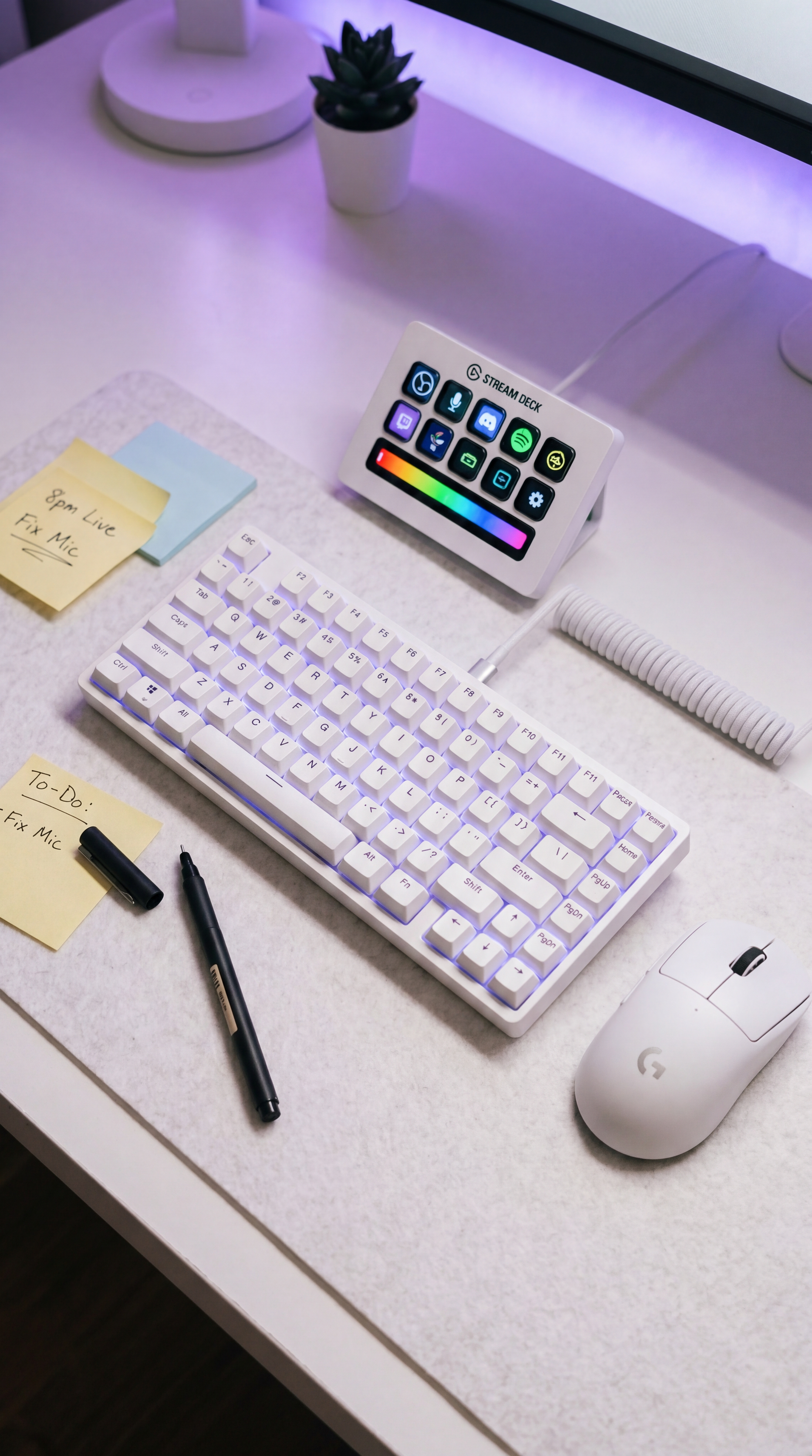 Overhead close-up of white TKL mechanical keyboard with coiled cable, white Elgato Stream Deck Plus, white Logitech mouse, sticky notes, and pens on felt desk mat
